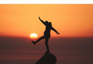 Silhouette of person balancing on one foot on a rock peak with sunset in background